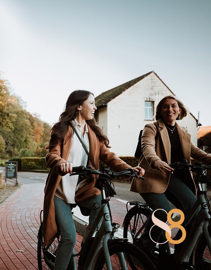 Twee dames Fietsen op een fietspad Langs het uithangbord van Brasserie De Groeve, langs een bosrand 