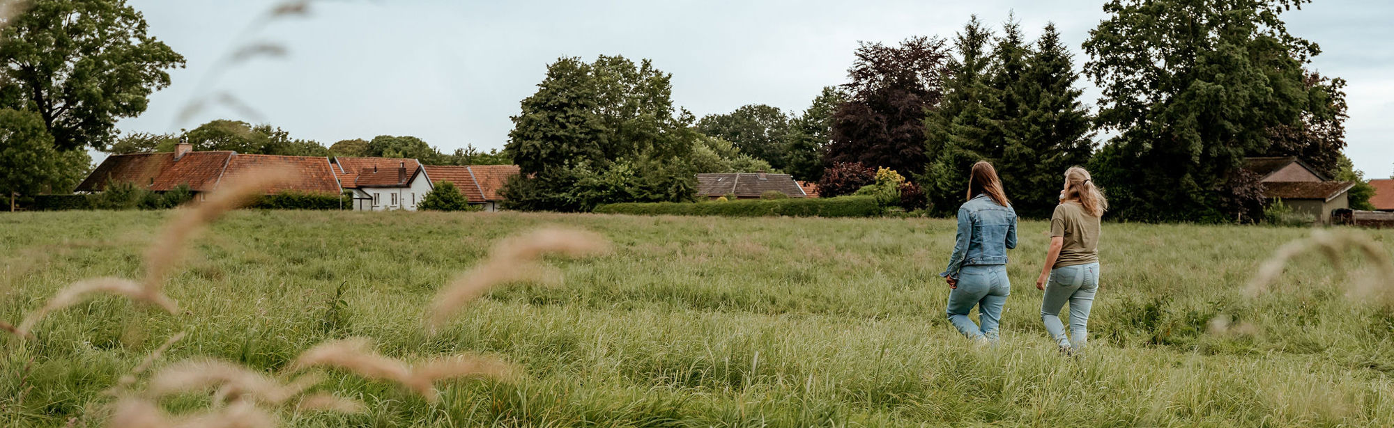 Twee dames wandelen door een weide met een boerderij op de achtergrond.