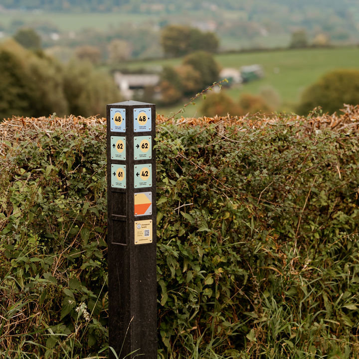 Knooppuntenpaal 48 staat tegen een heg en heeft op de achtergrond een uitzicht op het heuvellandschap op een bewolkte dag