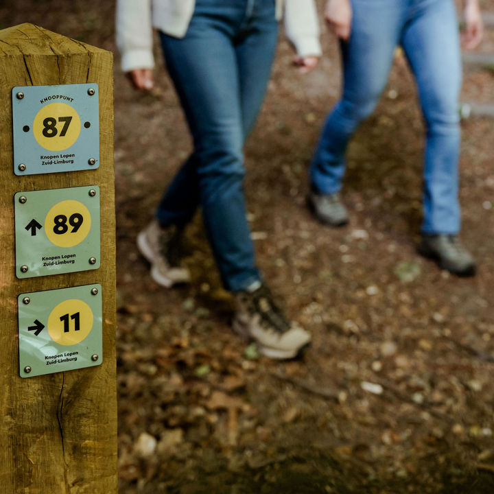 Een houten knooppuntenpaaltje in het bos met twee paar benen van vrouwen die over een onverhard pad wandelen