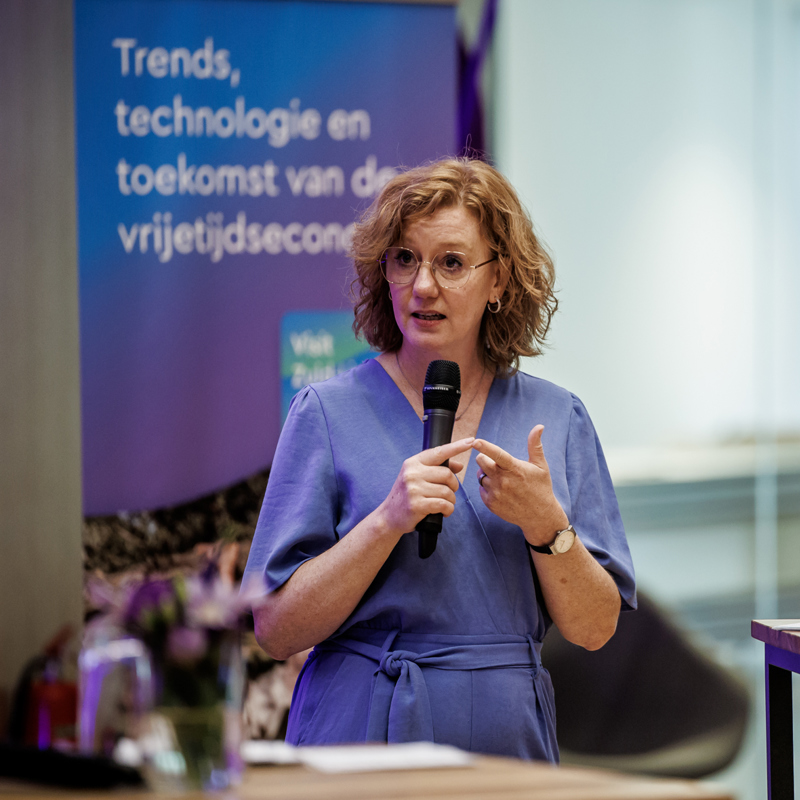 Manon spreekt op een congres van visit zuid-limburg met een microfoon in haar hand en een man op de achtergrond naast een statafel