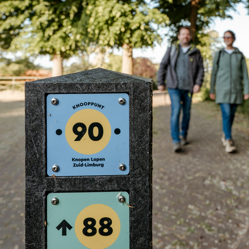 Knooppuntenpaaltje met twee wandelende mensen in de achtergrond